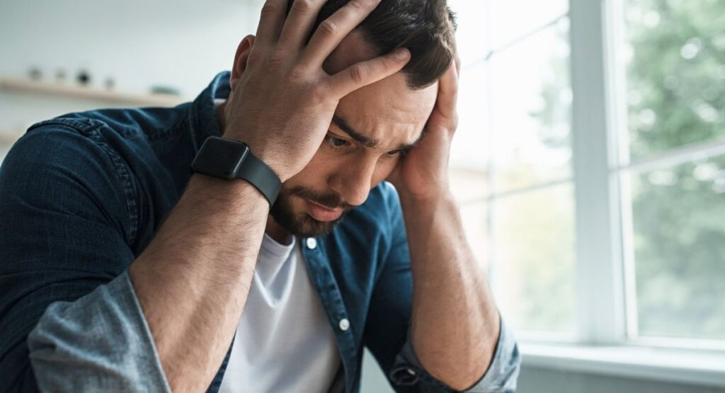 Homem com expressão de estresse, segurando a cabeça com as mãos, em ambiente interno com luz natural ao fundo. Ansiedade tem cura