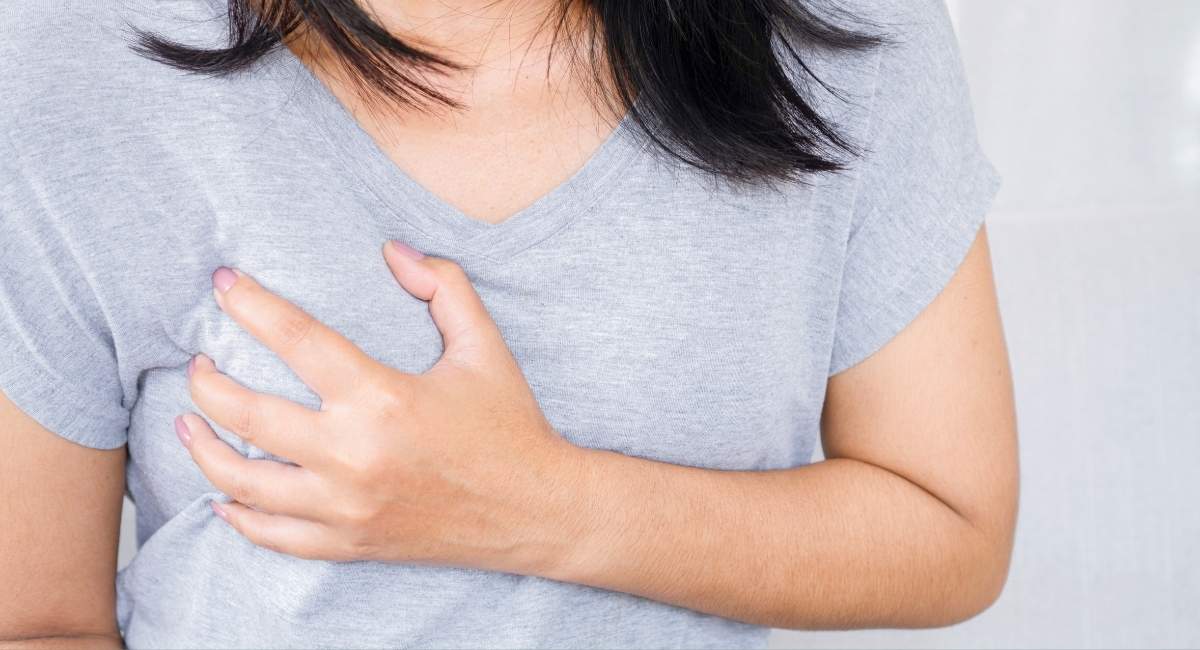 Mulher com a mão sobre o peito, em gesto que indica dor ou desconforto torácico, em fundo neutro.