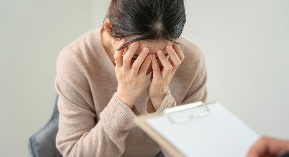 Qual o Impacto das Redes Sociais na Saúde Mental dos Jovens? 15 Mulher com as mãos no rosto, em gesto de angústia ou cansaço, durante uma consulta com profissional que segura uma prancheta.