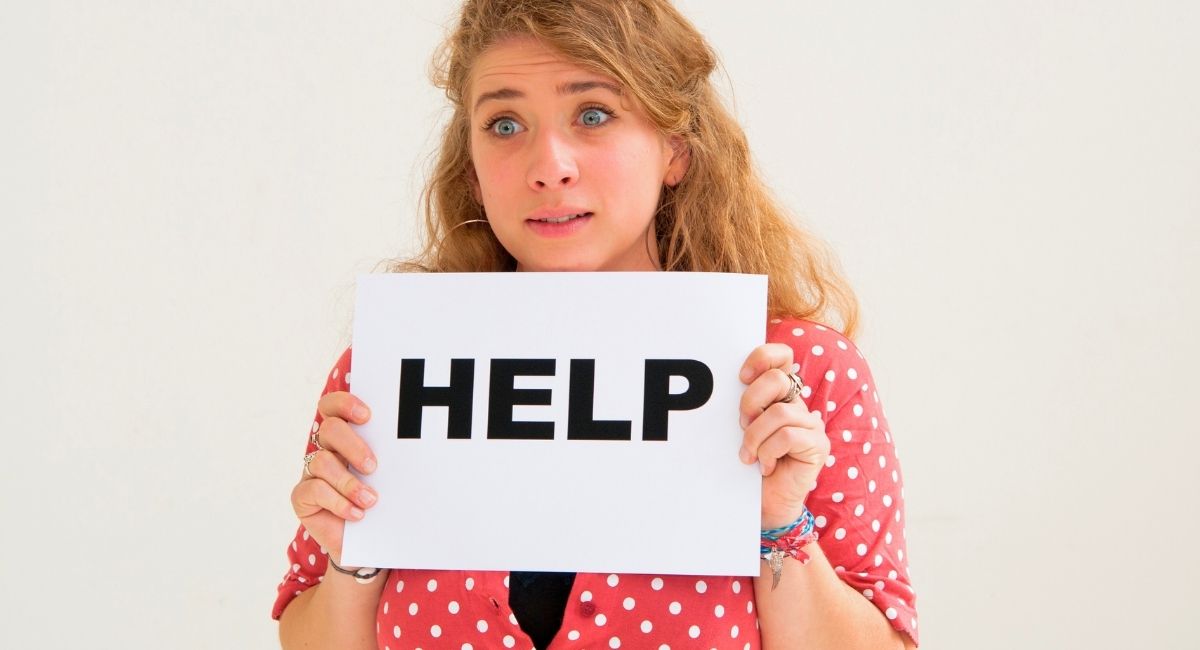 Mulher com expressão de preocupação segurando um cartaz com a palavra HELP, pedindo ajuda em fundo neutro.