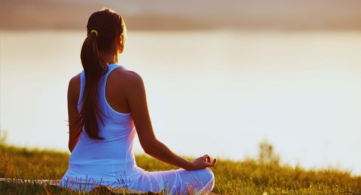 Mulher meditando em posição de lótus ao ar livre, com vista para um lago ao pôr do sol, em ambiente tranquilo e natural meditação