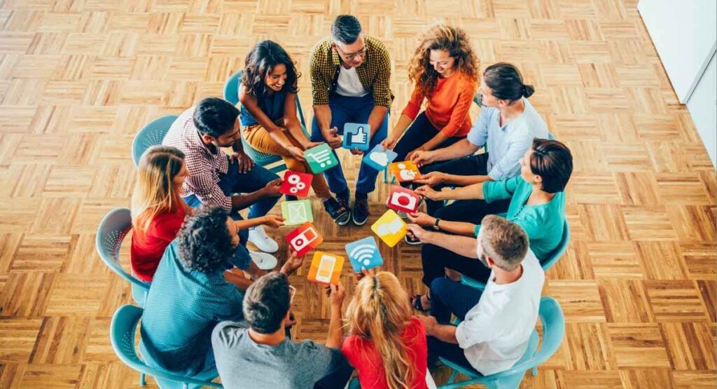 Grupo diverso de pessoas sentadas em círculo, compartilhando cartões com ícones de tecnologia, representando colaboração e inovação em equipe. Saúde Social