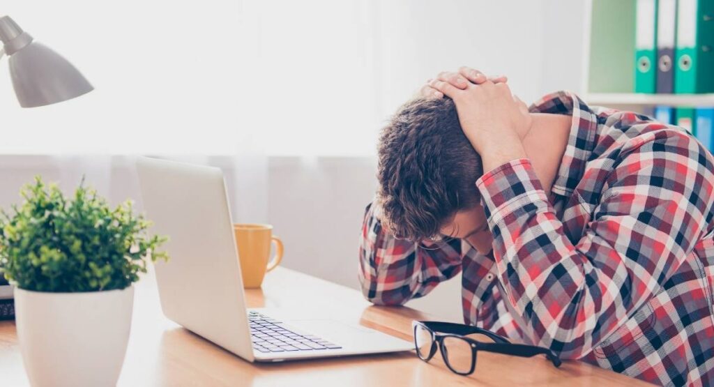 Homem com a cabeça nas mãos, sentado à mesa de trabalho com laptop, expressando estresse ou cansaço em ambiente doméstico. Ansiedade dá dor de cabeça