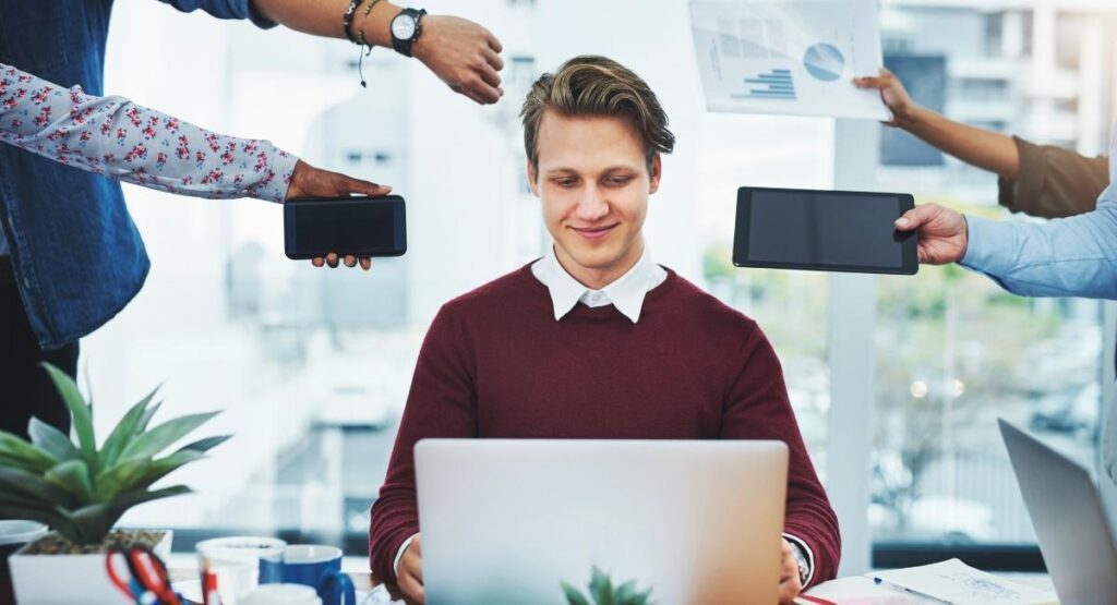 Homem trabalhando em laptop enquanto colegas oferecem dispositivos e documentos, representando colaboração e produtividade em ambiente de escritório Controle emocional
