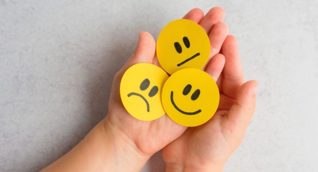 Mãos segurando três emojis amarelos com expressões de tristeza, neutralidade e felicidade, simbolizando diferentes estados emocionais. Saúde Emocional