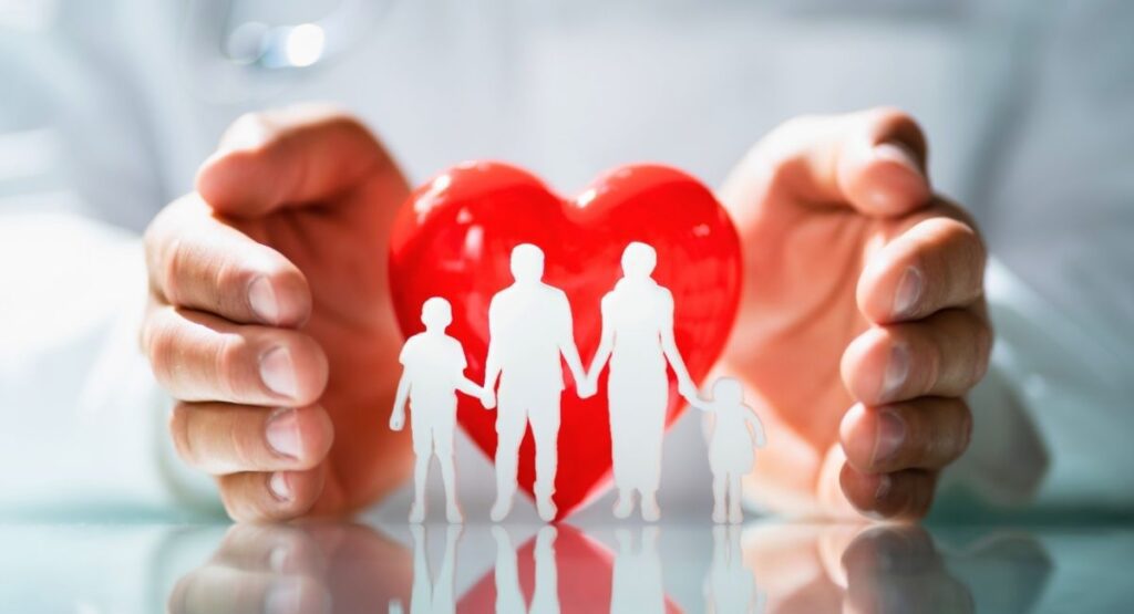 Mãos segurando um coração vermelho com silhuetas de uma família, simbolizando cuidado, proteção e bem estar familiar. Saúde Social