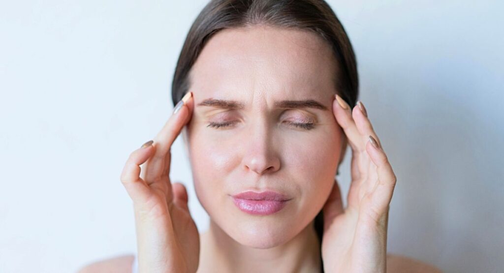 Mulher com os olhos fechados, pressionando suavemente as têmporas com os dedos, em gesto de relaxamento ou alívio de tensão, em fundo claro e neutro. Feridas na cabeça e ansiedade