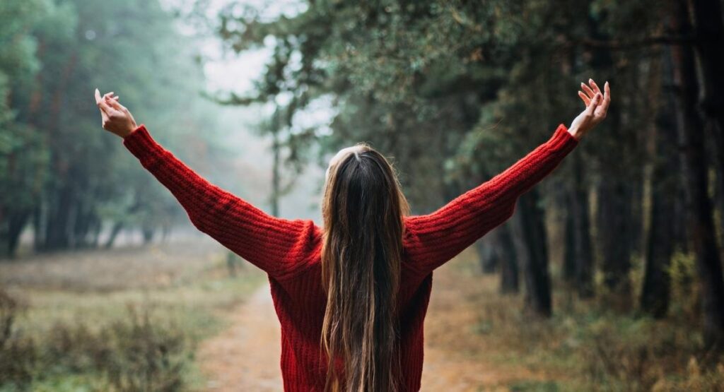 Mulher de costas, com os braços levantados em um bosque nebuloso, vestindo suéter vermelho, expressando liberdade e conexão com a natureza. Saúde Emocional