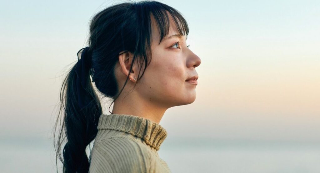 Mulher em perfil, olhando para o horizonte ao entardecer, com expressão serena e cabelos presos em rabo de cavalo, em ambiente calmo junto ao mar.