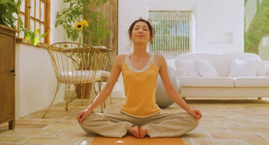 Mulher em posição de meditação no chão, com os olhos fechados e expressão serena, em um ambiente acolhedor e iluminado por luz natural meditação