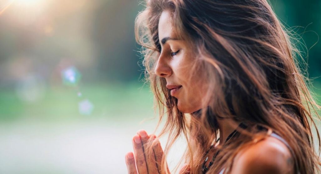 Mulher em posição de meditação ou oração, com olhos fechados e mãos unidas, em ambiente externo com fundo verde e luz suave Meditação IASD