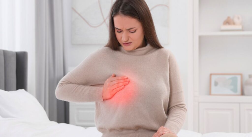 Mulher segurando o peito com expressão de dor, com área destacada em vermelho, indicando desconforto ou sintoma cardíaco em ambiente doméstico. Ansiedade causa dor na mama
