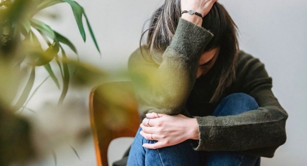 Mulher sentada com a cabeça baixa, abraçando os joelhos, em gesto de tristeza ou angústia, em ambiente simples e com planta ao fundo. Inteligência Emocional