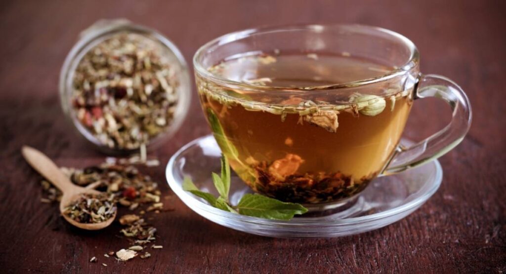 Xícara de chá de ervas transparente, com folhas e flores flutuando, ao lado de uma colher e pote com mistura seca, em fundo rústico e acolhedor. Calmante natural para ansiedade