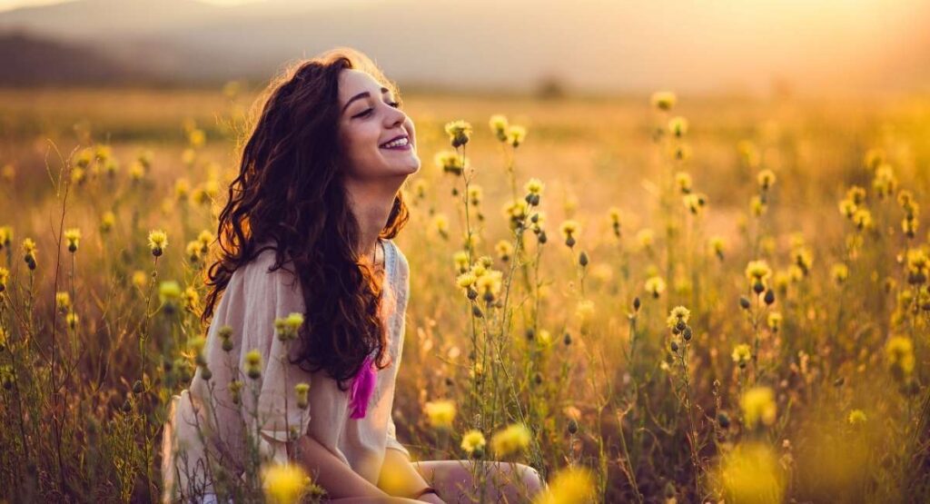 uma mulher sorridente, de cabelos cacheados, sentada em um campo de flores amarelas durante o pôr do sol. A luz suave do entardecer cria um clima sereno e natural. Meditação IASD